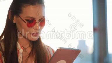 年轻迷人的女士戴着红色太阳镜，在咖啡馆里使用带有触摸屏的平板电脑。 机场或机场的美女